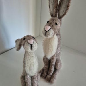 Puede incluir: Dos figuritas de conejo de fieltro marrón y blanco. El conejo más grande está sentado erguido con las orejas levantadas. El conejo más pequeño está sentado con la cabeza inclinada. Ambos conejos tienen ojos negros y narices rosas.