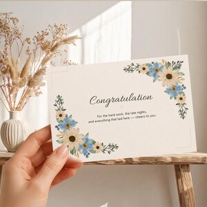 Peut inclure: Une carte de vœux rectangulaire blanche ornée d'une bordure florale de fleurs bleues et crème dans les coins. Le texte indique : Congratulation. For the hard work, the late nights, and everything that led here — cheers to you.