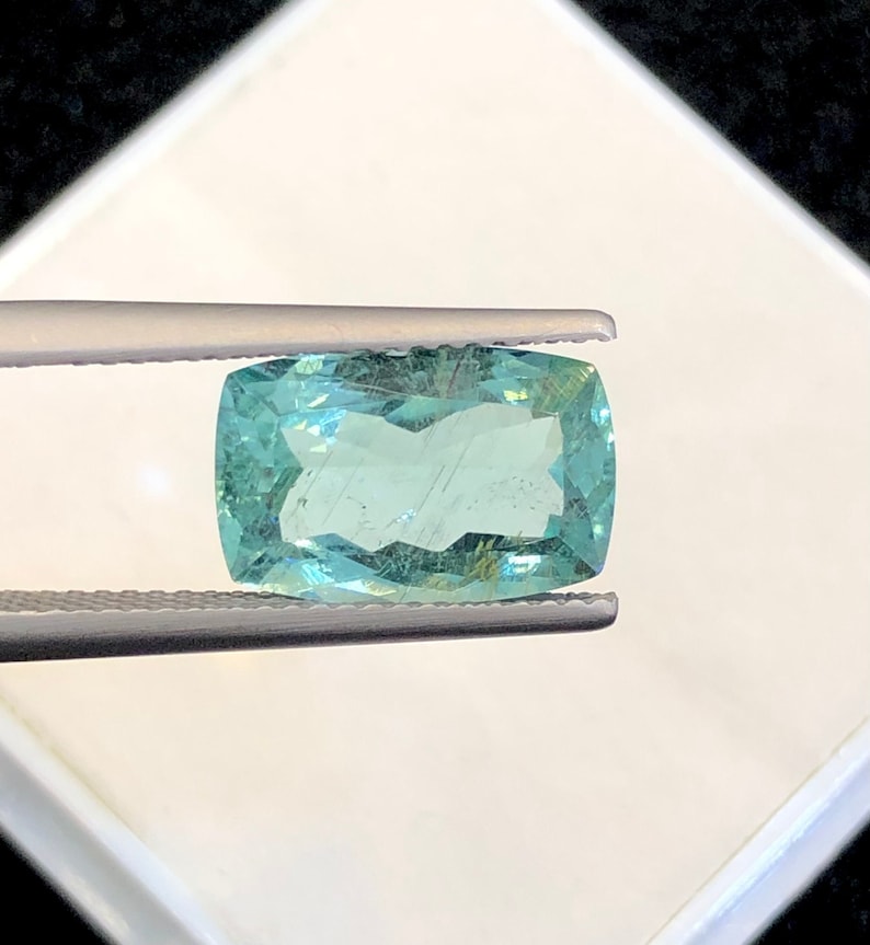 May include: A light blue, rectangular, faceted gemstone held by tweezers. The gemstone is transparent and has a slight green hue.