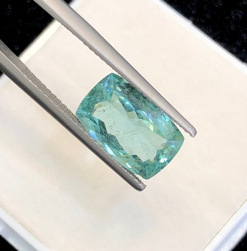 May include: A light green gemstone with a rectangular shape, held by tweezers against a white background.