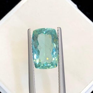 May include: A light blue gemstone with a rectangular shape, held in a pair of tweezers. The gemstone is clear and has a slight green tint.