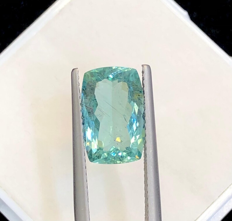 May include: A light blue gemstone with a rectangular shape, held in a pair of tweezers. The gemstone is clear and has a slight green tint.