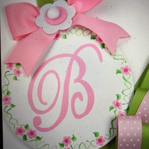 May include: A white circular wooden sign with a pink floral border and a large pink letter "B" in the center. The sign is decorated with a pink bow and a white flower with a pink center.