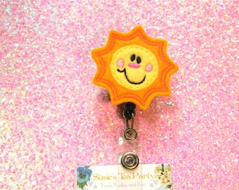 May include: A yellow felt sun with a smiling face and pink cheeks, attached to a black and silver retractable badge holder. The sun is on a pink glitter background.