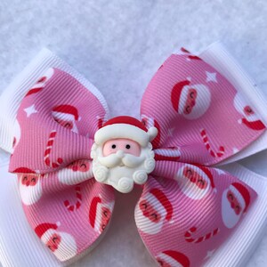 May include: Pink Christmas bow with a white Santa Claus face in the center. The bow is made of ribbon with a repeating pattern of Santa Claus faces and candy canes.
