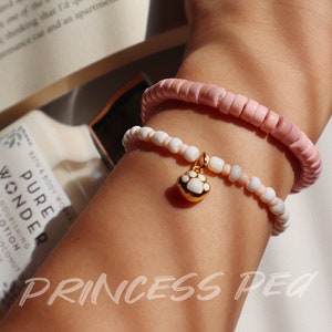 May include: Two beaded bracelets on a wrist. The top bracelet is pink and the bottom bracelet is white with a gold paw print charm.