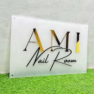 Op de afbeelding: Een wit bord met gouden letters waarop in een cursief schrift "AMI Nail Room" staat.