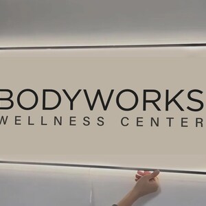 May include: A rectangular, wall-mounted sign with a light-colored background and a black frame. The text reads BODYWORKS WELLNESS CENTER in bold, black, sans-serif capital letters. The sign features integrated perimeter LED backlighting.