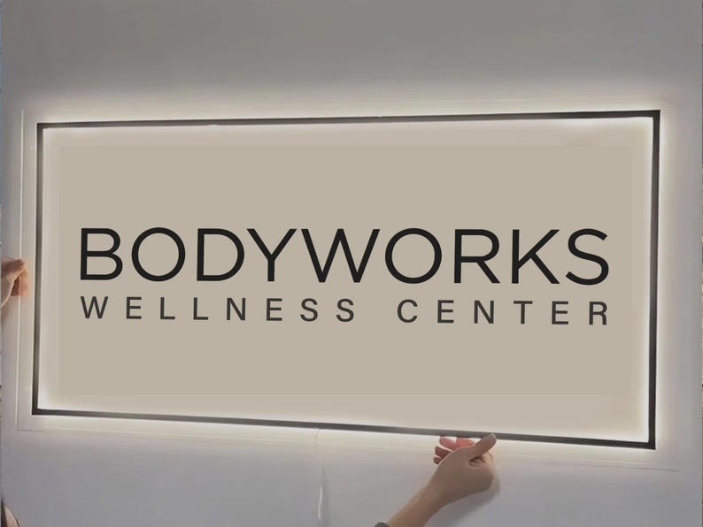 May include: A rectangular, wall-mounted sign with a light-colored background and a black frame. The text reads BODYWORKS WELLNESS CENTER in bold, black, sans-serif capital letters. The sign features integrated perimeter LED backlighting.