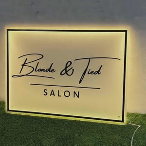 May include: A rectangular, backlit salon sign with a white face and a black border. The text reads "Blonde & Tied" in a black cursive script, with the word "SALON" in black capital letters underneath. The sign emits a warm, glowing light.