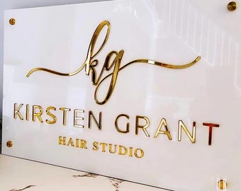 May include: White acrylic sign with gold lettering. The sign reads "kg" in a cursive font, followed by "Kirsten Grant Hair Studio".