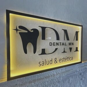 May include: A rectangular wall sign with a black frame and backlit yellow glow. It features a black tooth icon with a star, the large letters DM, the text DENTAL MN, and the smaller text salud & est&eacute;tica in a black sans-serif font.