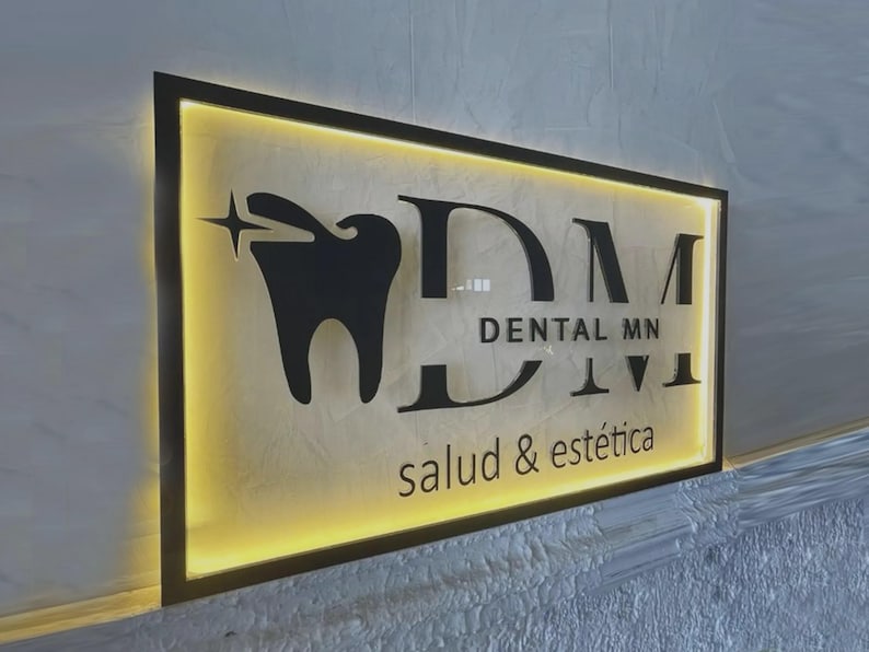 May include: A rectangular wall sign with a black frame and backlit yellow glow. It features a black tooth icon with a star, the large letters DM, the text DENTAL MN, and the smaller text salud & est&eacute;tica in a black sans-serif font.