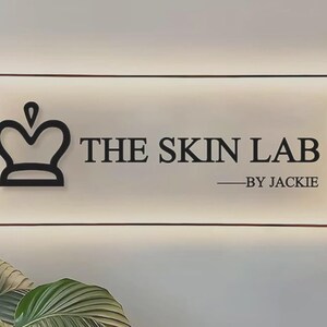 May include: A rectangular, backlit acrylic sign featuring a black crown icon on the left and the black text THE SKIN LAB BY JACKIE on the right. The sign is mounted with four metallic corner standoffs and emits a soft glow from behind.