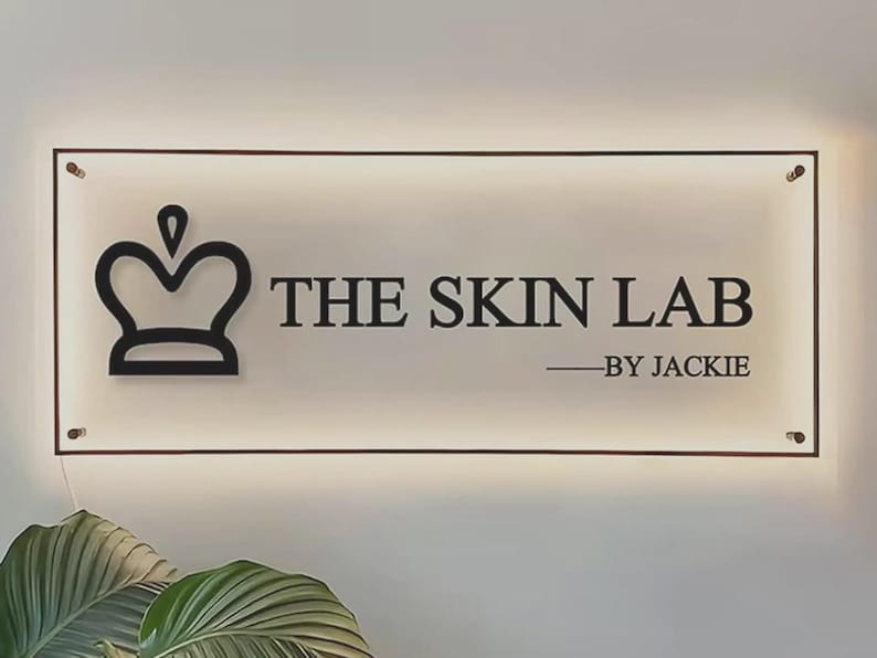 May include: A rectangular, backlit acrylic sign featuring a black crown icon on the left and the black text THE SKIN LAB BY JACKIE on the right. The sign is mounted with four metallic corner standoffs and emits a soft glow from behind.