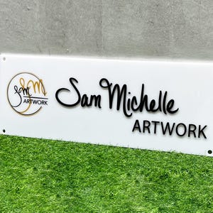 May include: White rectangular sign with black text that reads "Sam Michelle Artwork" and a gold logo with the text "Sam Michelle Artwork".
