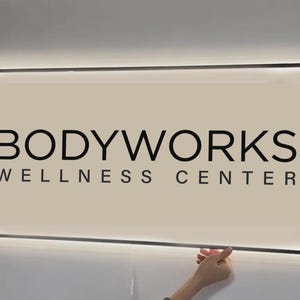 May include: A rectangular, wall-mounted sign with a light beige background and a thin black border. The text "BODYWORKS" appears in large, bold, black sans-serif capital letters, with "WELLNESS CENTER" in smaller black letters below it.