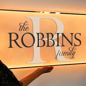 Puede incluir: Un letrero blanco iluminado con un contorno negro y una gran letra blanca "R" en el centro. El texto "the ROBBINS family" está escrito en letras negras en el letrero.