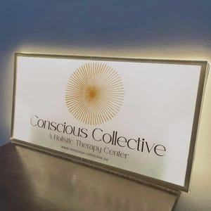 May include: A rectangular, illuminated sign with a white face and a gold sunburst graphic. The text reads Conscious Collective, A Holistic Therapy Center, and www.conscious-collective.co in black lettering. The sign emits a soft glow from its edges.