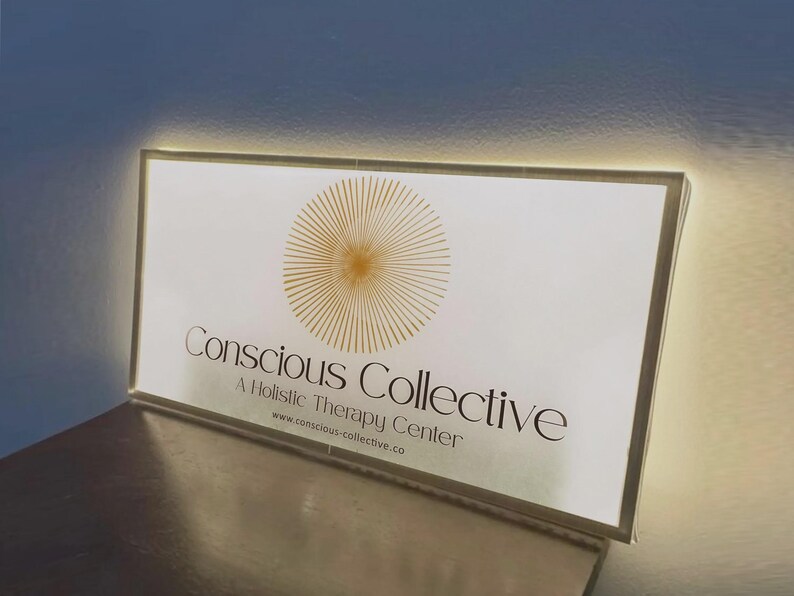 May include: A rectangular, illuminated sign with a white face and a gold sunburst graphic. The text reads Conscious Collective, A Holistic Therapy Center, and www.conscious-collective.co in black lettering. The sign emits a soft glow from its edges.