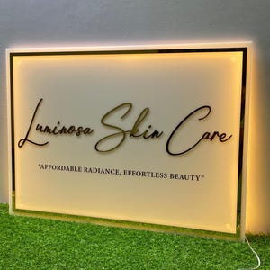 Pode incluir: Letreiro luminoso para Luminousa Skin Care. O letreiro apresenta o nome da marca em escrita elegante, com o slogan «Affordable Radiance, Effortless Beauty». O letreiro tem um fundo branco com uma borda dourada e preta e é iluminado com um brilho quente.