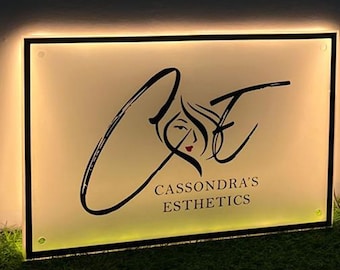 May include: Illuminated sign with the text "Cassondra's Esthetics". The sign features a stylized logo with a woman's face and flowing hair. The sign has a black border and is lit with a warm, inviting glow.