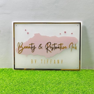 Op de afbeelding: Een rechthoekig bord met een witte achtergrond, gouden rand en de woorden "Beauty & Restorative Ink" in gouden letters. Een roze vorm staat achter de tekst, met de woorden "BY TIFFANY" eronder. Kleine roze accenten staan boven de tekst.