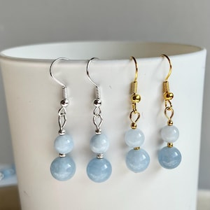 May include: Four pairs of dangle earrings with light blue gemstone beads. Two pairs have silver-coloured hooks and chains, while the other two feature gold-coloured hooks and chains. The earrings are displayed against a white background.