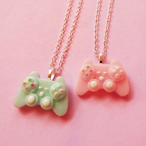 May include: Two silver chain necklaces with pastel-colored game controller pendants. The pendant on the left is light blue with white and multi-colored details. The pendant on the right is pink with white and multi-colored details.