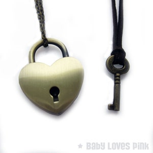 May include: A heart-shaped padlock necklace with a key on a brown cord. The padlock is made of gold-toned metal and has a keyhole in the center. The key is also gold-toned metal.