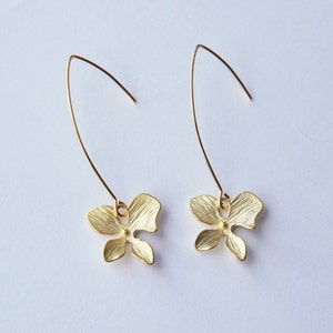 May include: A pair of gold-tone dangle earrings featuring a delicate flower design. The earrings have a long, thin wire that hangs from the flower.
