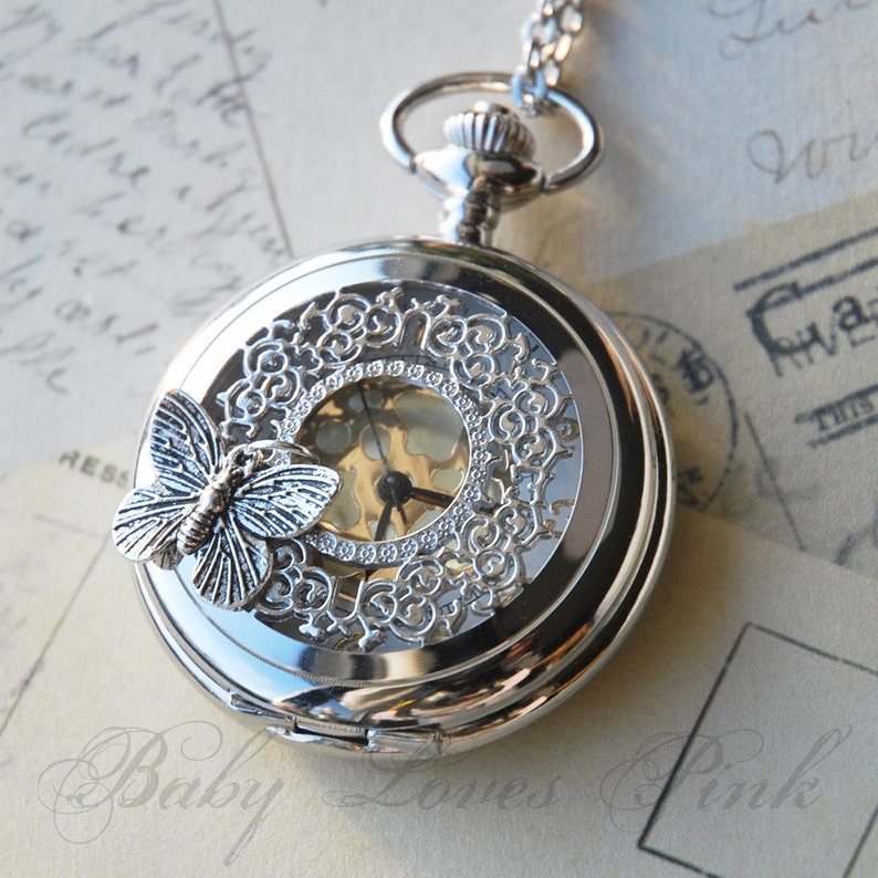 May include: Silver pocket watch with a delicate filigree design and a butterfly charm. The watch face is visible through the openwork design.