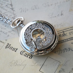 May include: Silver pocket watch with a delicate butterfly design and a clear face. The watch has a filigree design around the edge and a chain attached.