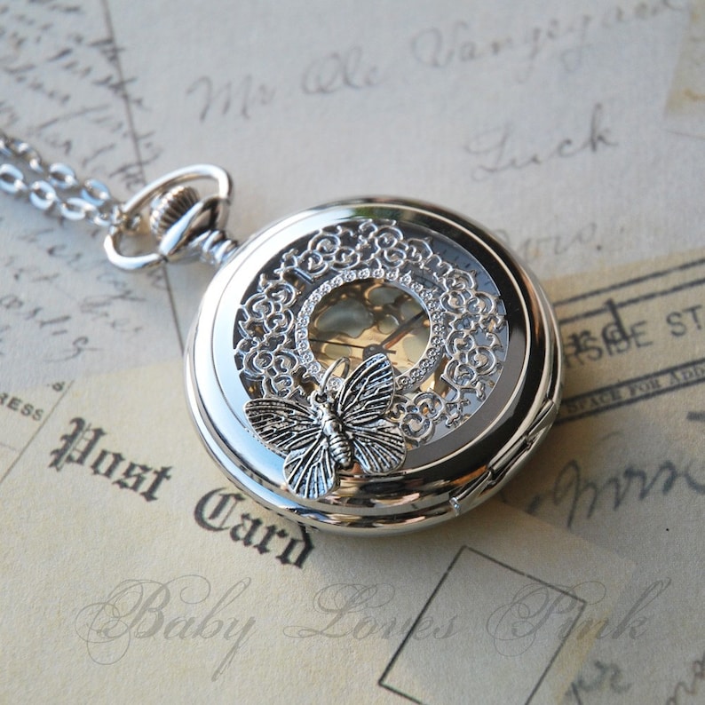 May include: Silver pocket watch with a delicate butterfly design and a clear face. The watch has a filigree design around the edge and a chain attached.