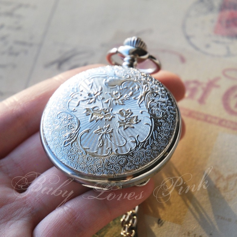 May include: Silver pocket watch with an ornate floral design on the front. The watch has a chain attached.