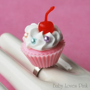 以下が含まれることがあります： 白いフロスティングと赤いチェリーがトッピングされたピンクのカップケーキ。カップケーキはカラフルなスプリンクルで飾られており、白い表面の上に置かれています。