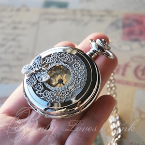 May include: Silver pocket watch with a delicate butterfly design and a clear face revealing the inner workings of the watch.