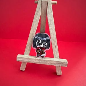 Könnte beinhalten: Ein kleiner, pixeliger Charakter-Pin, der eine Figur mit schwarzen Haaren, einem roten Schal und einem braunen Outfit zeigt. Der Pin wird auf einer Miniatur-Holzstaffelei vor rotem Hintergrund präsentiert. Der Pin ist etwa 5 cm hoch.