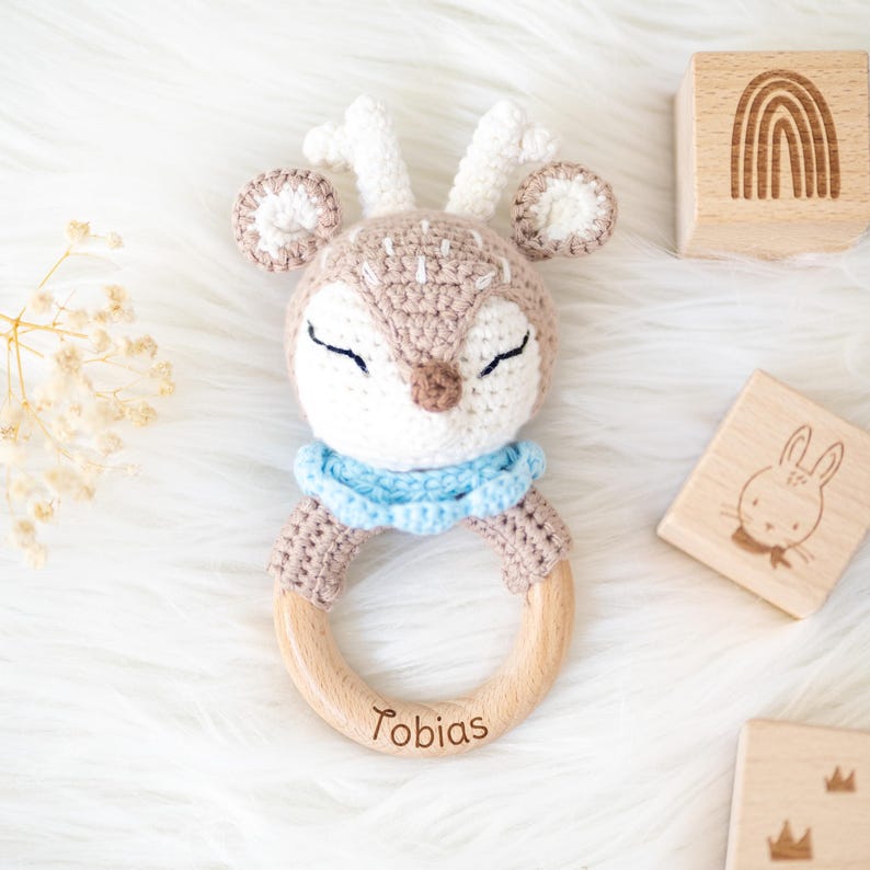 May include: A crocheted deer rattle with a wooden ring, personalized with the name "Tobias". The rattle is brown and white with blue accents.