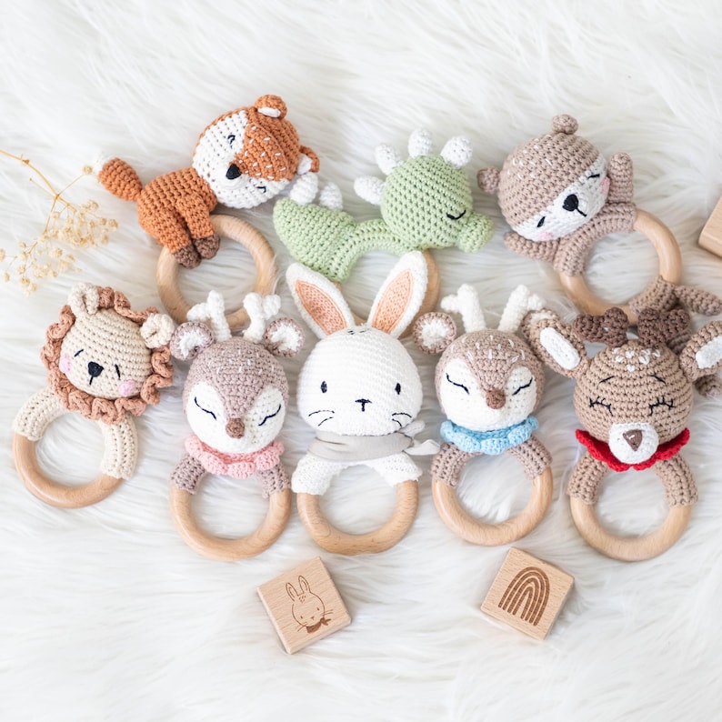 May include: A set of nine crocheted animal teethers with wooden rings. The animals include a fox, a dinosaur, a deer, a rabbit, a lion, and a bear. The teethers are in various colors, including brown, green, white, and red.
