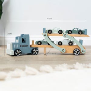May include: A blue wooden toy truck with the name "Fabian" on the side, carrying four light green toy cars on a double-decker trailer. The truck is 13 inches long, 8 inches wide, and 38 inches tall.