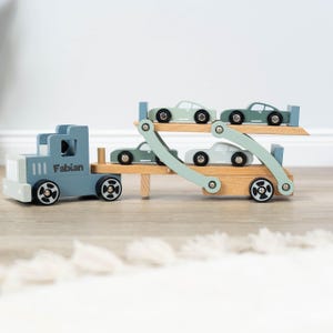 May include: A blue wooden toy truck with the name "Fabian" on the side, carrying four smaller green wooden toy cars on a wooden ramp.