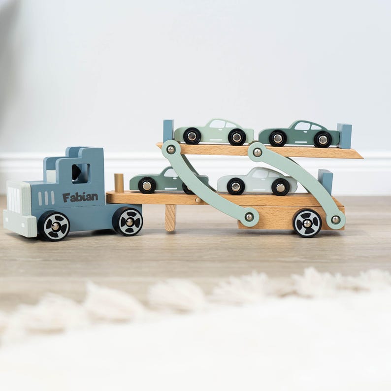 May include: A blue wooden toy truck with the name "Fabian" on the side, carrying four smaller green wooden toy cars on a wooden ramp.