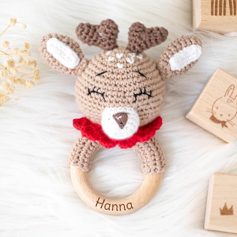May include: A crocheted reindeer rattle with a wooden ring, personalized with the name "Hanna". The reindeer has a red and white ruffle collar and closed eyes.