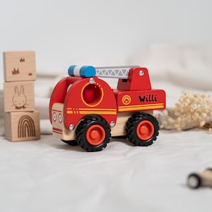 Könnte beinhalten: Rotes Holzspielzeug-Feuerwehrauto mit einer Leiter, schwarzen Rädern und einem blauen Licht oben. Das Auto hat einen gelben Streifen und den Namen "Willi" an der Seite.