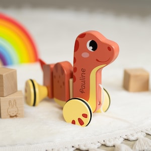 Könnte beinhalten: Ein hölzernes Nachziehtier in Form eines Dinosauriers in Orange und Gelb, mit dem Namen "Pauline" auf der Seite. Das Spielzeug hat ein lächelndes Gesicht, rote Punkte und gelbe Räder. Ein Regenbogen und Holzblöcke sind im Hintergrund.
