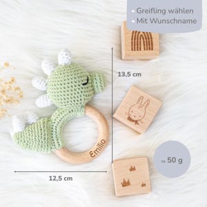 May include: Green crocheted dinosaur rattle with a wooden ring, personalized with the name "Emilio". The rattle is 12.5 inches long and comes with three wooden blocks, each with a different design: a rainbow, a bunny, and a crown. The blocks are 13.5 cm wide and the set weighs approximately 50 grams.
