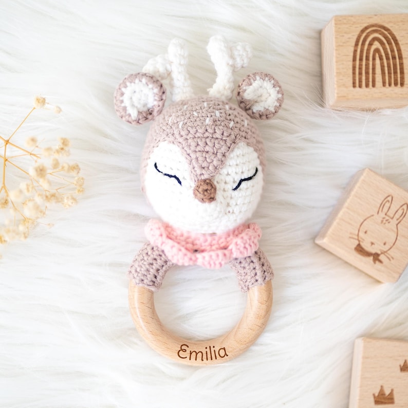 May include: A crocheted deer rattle with a pink ruffle and a wooden ring. The ring is engraved with the name "Emilia".