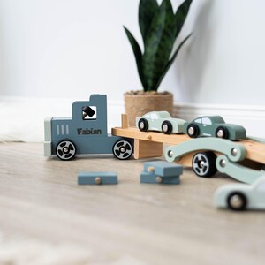 May include: A blue toy truck with the name "Fabian" on the side is carrying three smaller toy cars on a wooden platform. The cars are green and light blue. The truck and cars are made of wood and have wheels that roll.