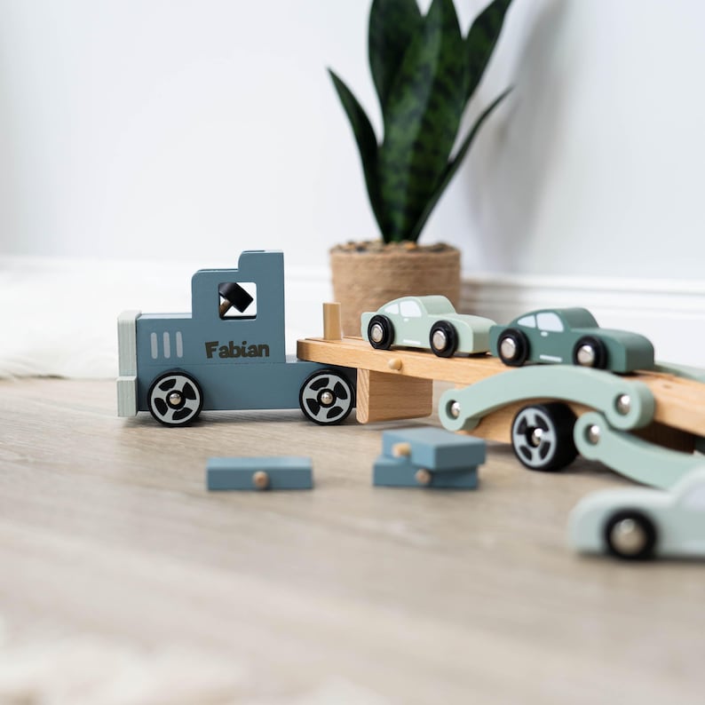 May include: A blue toy truck with the name "Fabian" on the side is carrying three smaller toy cars on a wooden platform. The cars are green and light blue. The truck and cars are made of wood and have wheels that roll.